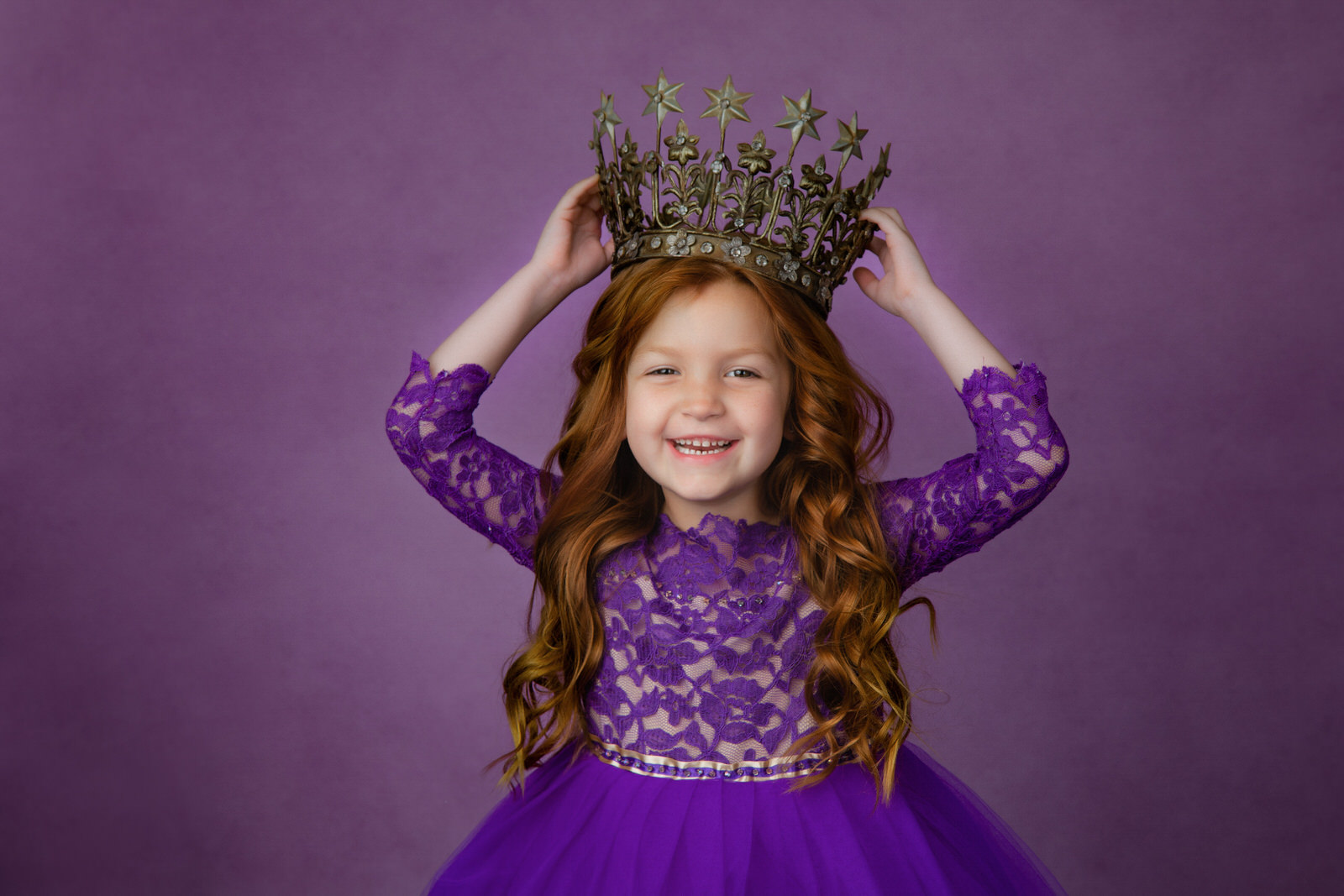 A young girl in a purple dress holds a large crown above her head in a studio DFW Kids fine art princess photography