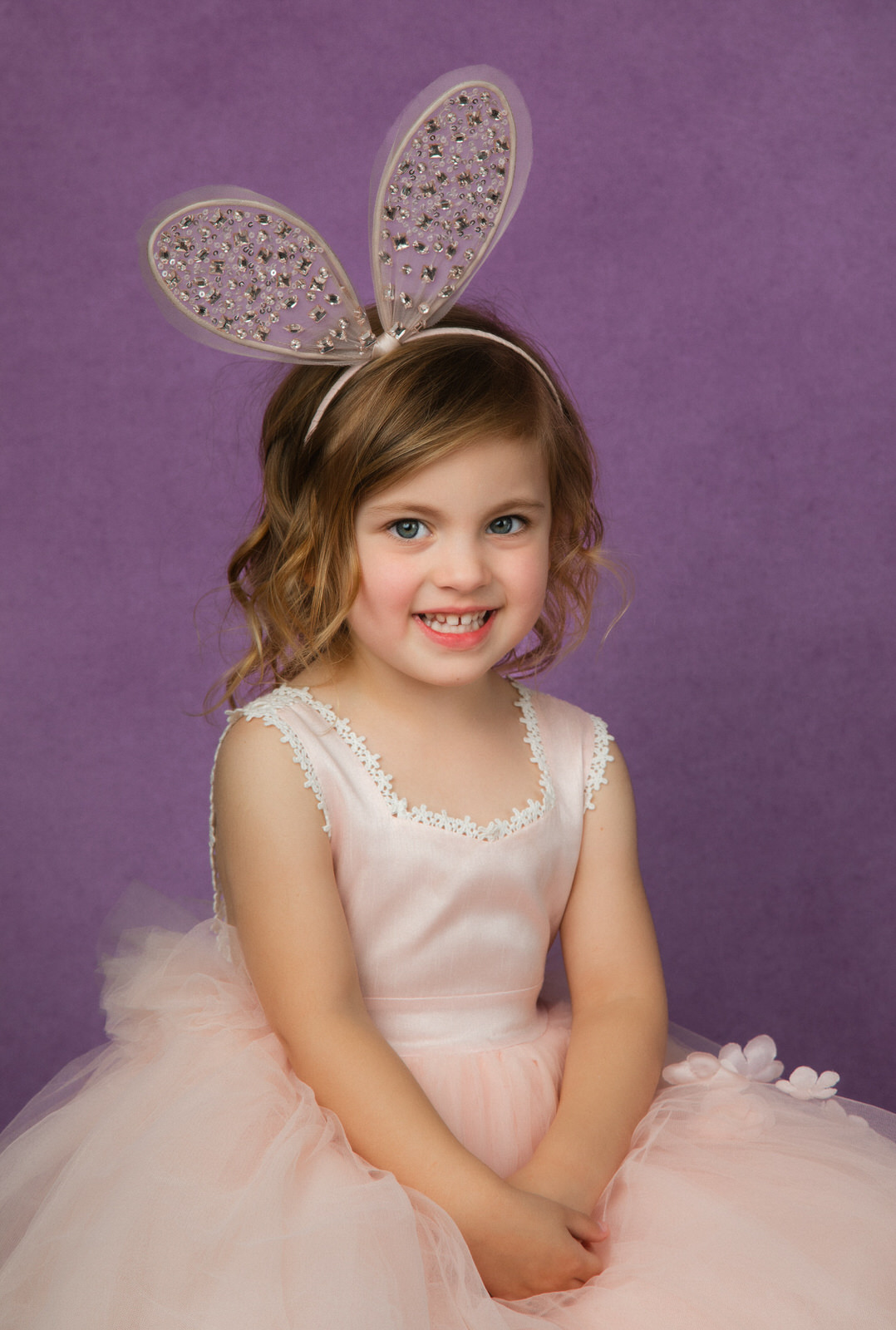 A young girl in a pink tutu and bunny ears headband sits on a stool in a studio DFW Kids