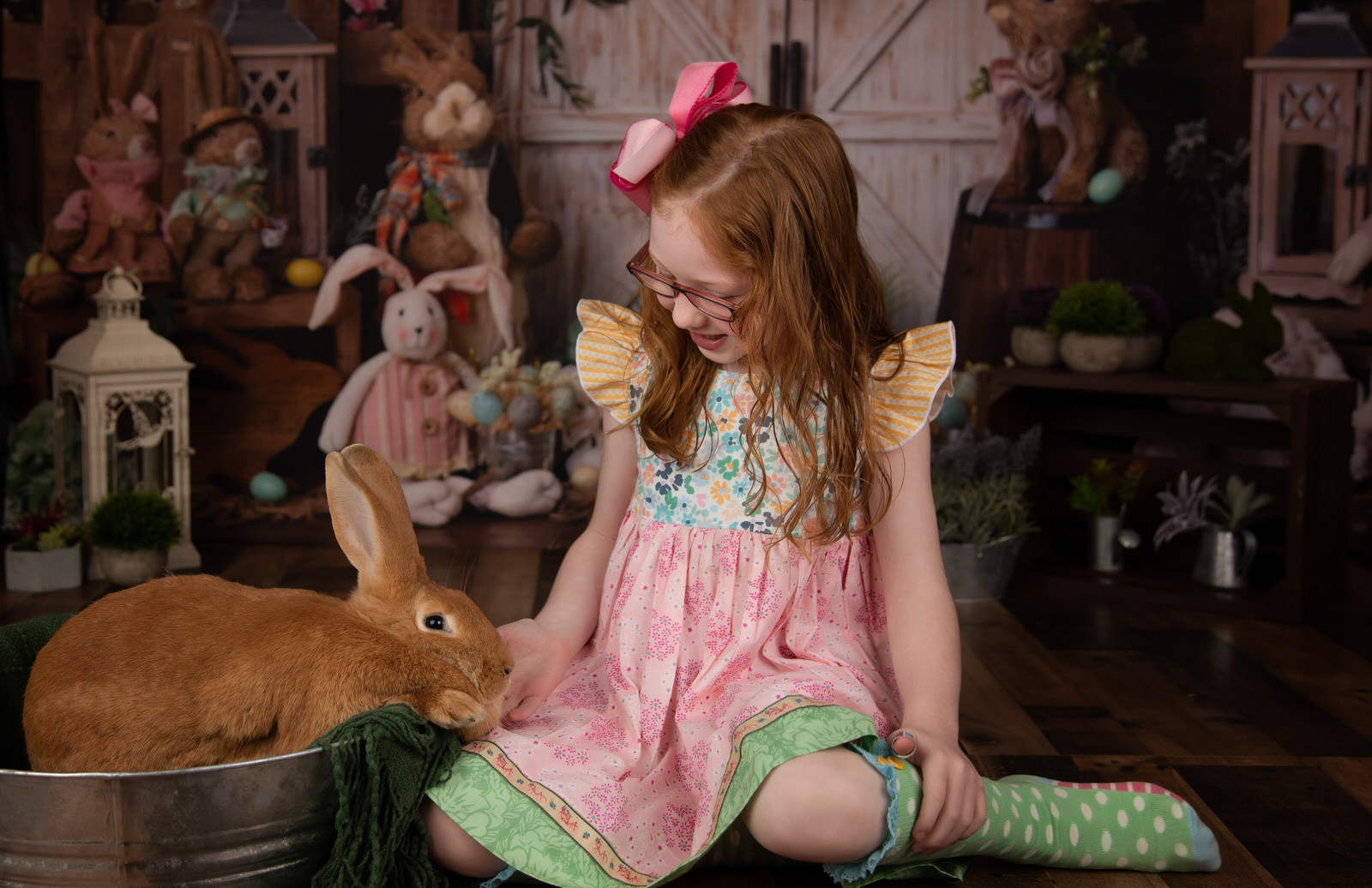 A young girl in a colorful dress and socks pets a bunny in a tin bucket