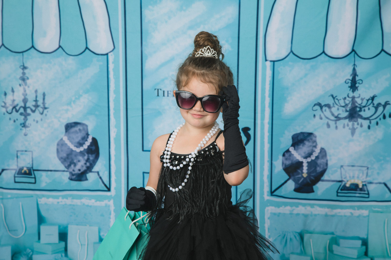 A young girl in a black flapper dress and large sunglasses holds a shopping bag in a studio