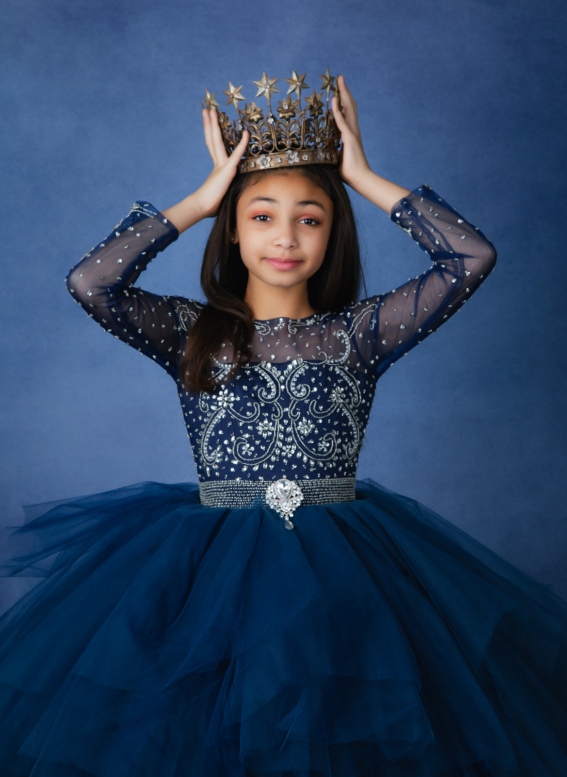 A young girl in an ornate blue dress holds a crown above her head in a studio dallas babysitters