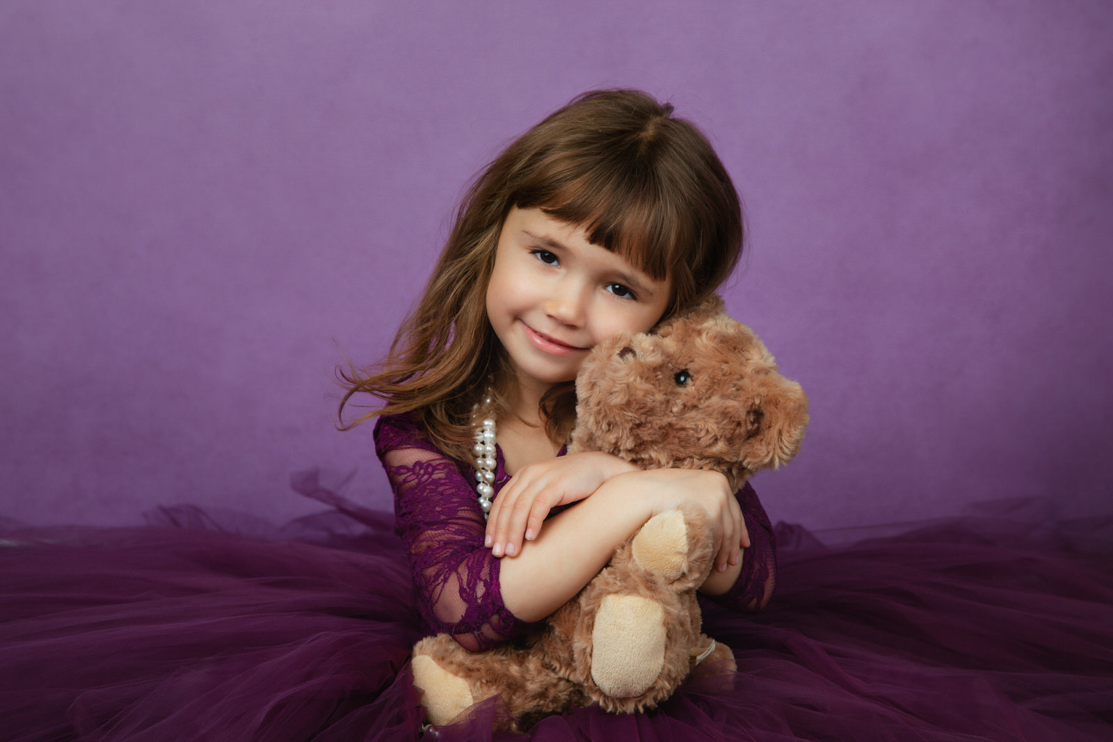 A young girl in a purple dress hugs onto a teddy bear in a studio