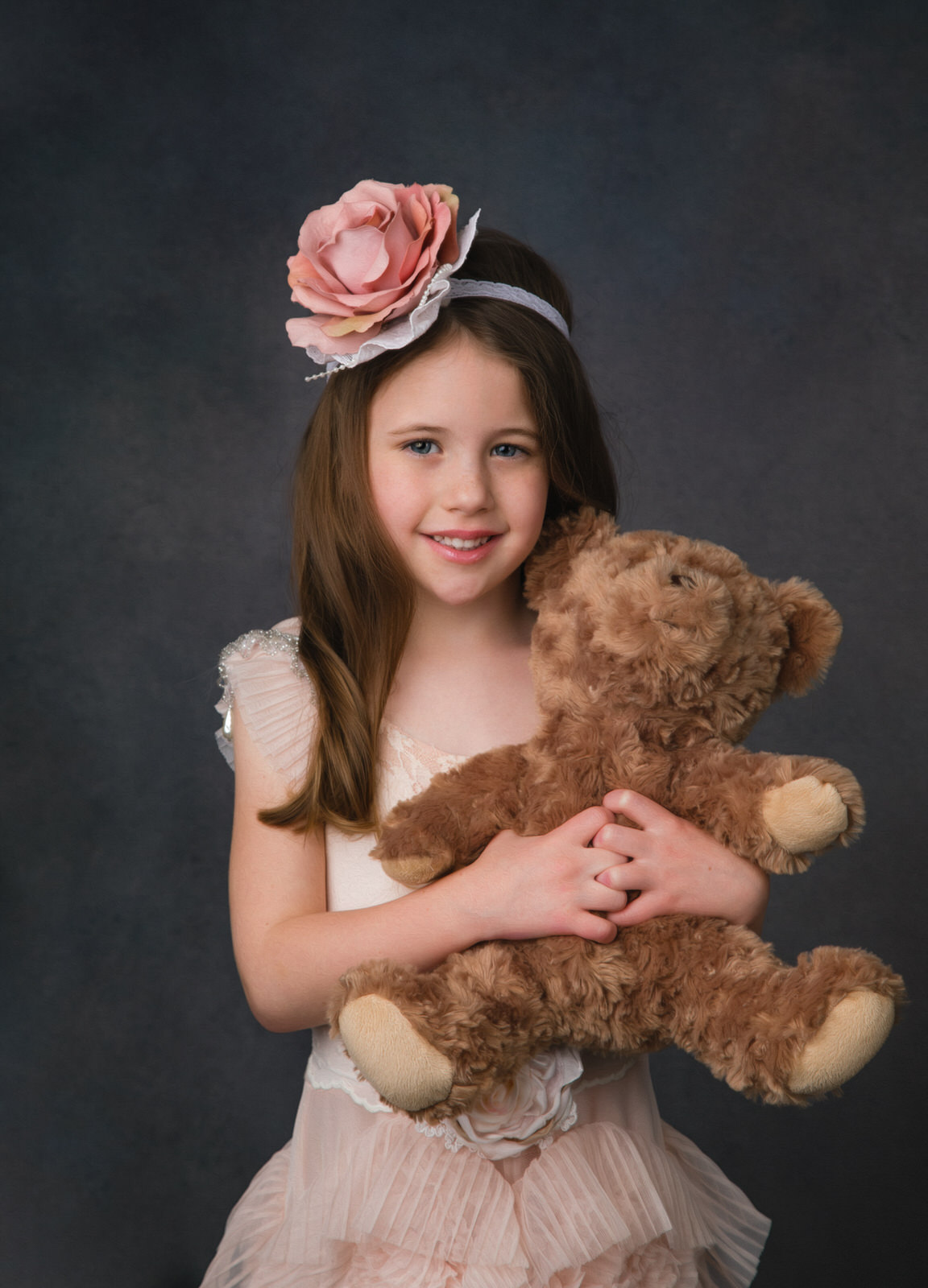 A young girl in a dress and large flower headband holds a brown teddy bear in a studio dallas pediatricians