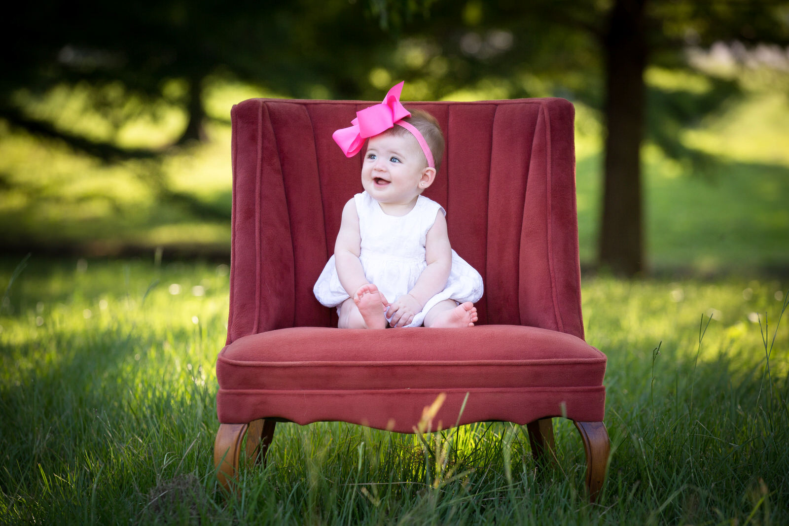 A young toddler in a white dress and large pink bow sits in a red chair in a park fort worth babysitters