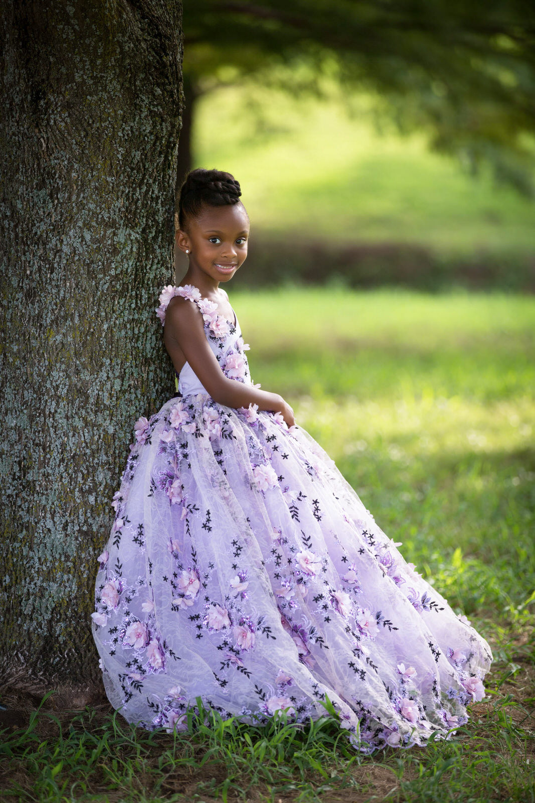 A young girl in a purple floral tulle dress leans against a large tree trunk fort worth babysitters