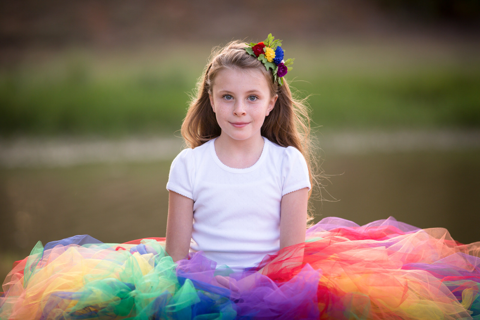 A young girl sits in a park in a multi-color tule dress and white shirt