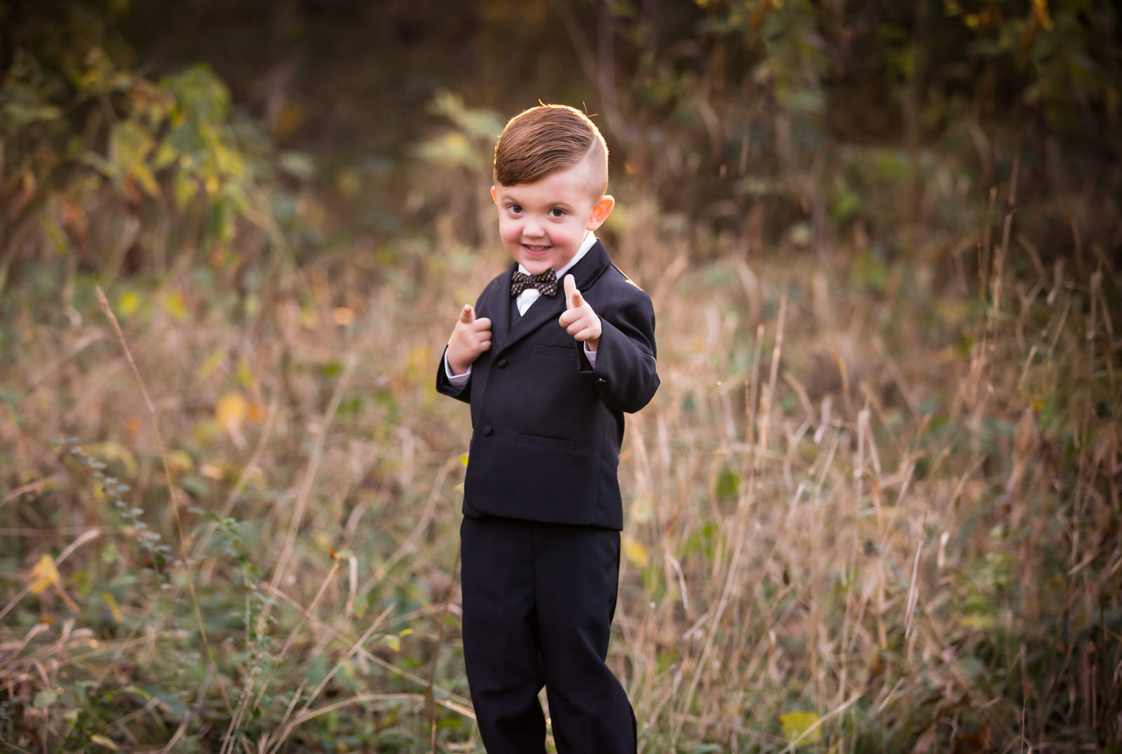 A young boy in a full suit and bowtie stands in a field of tall grass fort worth kids