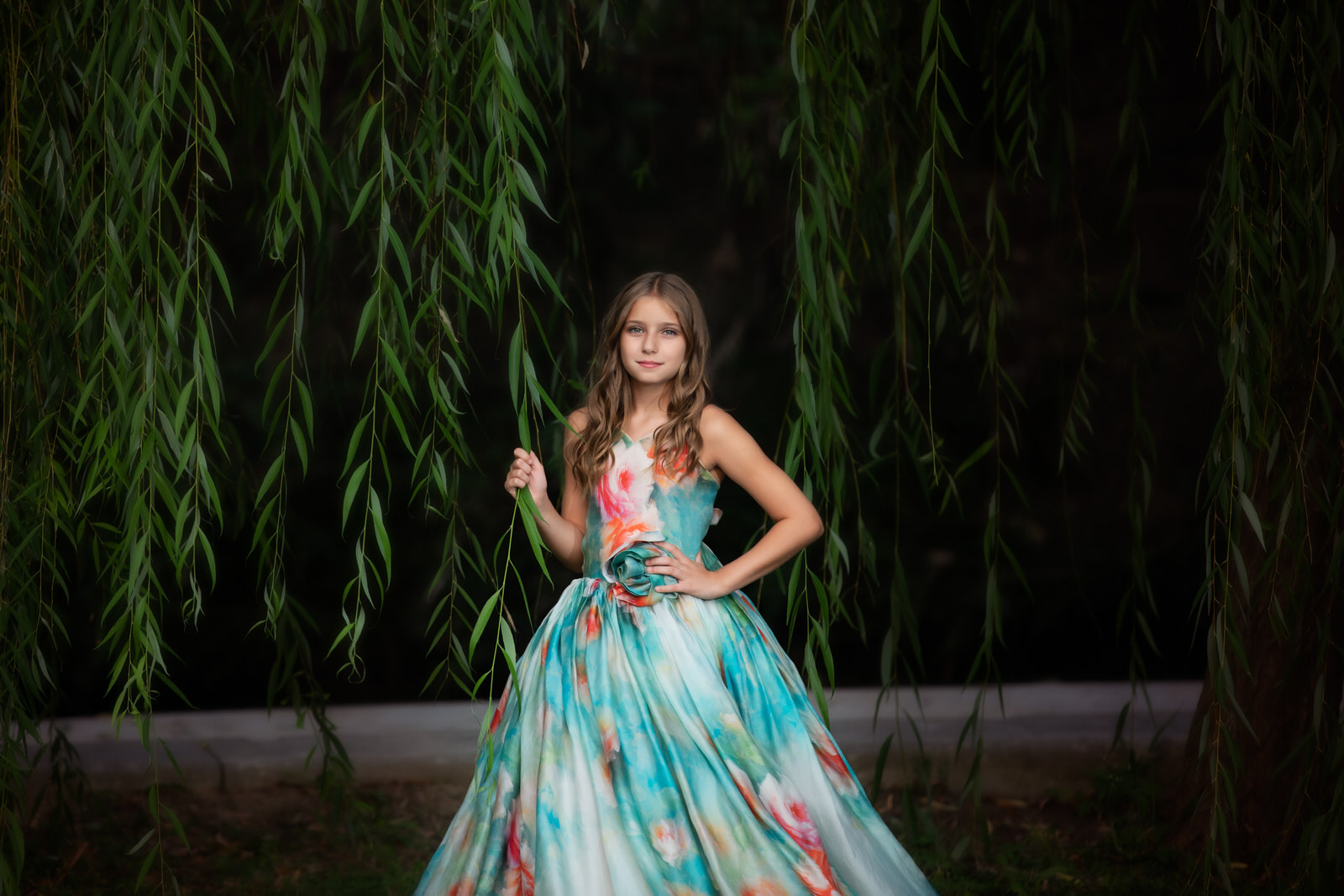 A young girl hold onto the branch of a large willow tree in a blue floral dress