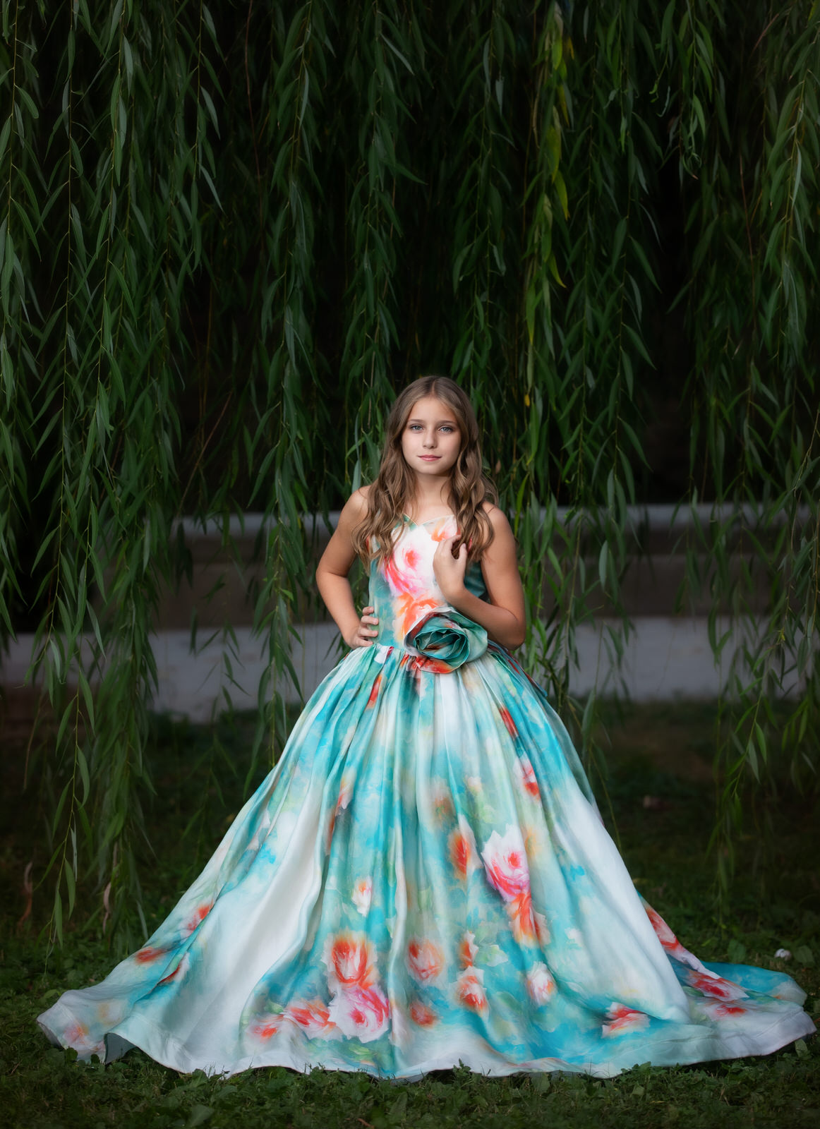 A teen girl stands under a willow tree in a large floral gown with a hand on her hip fort worth pediatricians