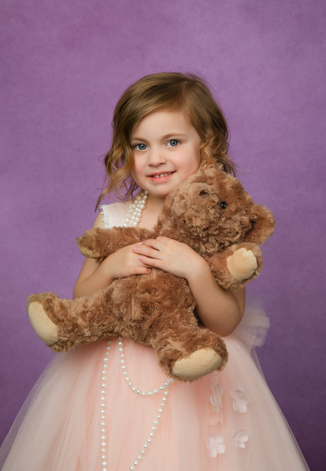 A young girl in a pink tule dress hugs a brown teddy bear in a studio fort worth toy stores