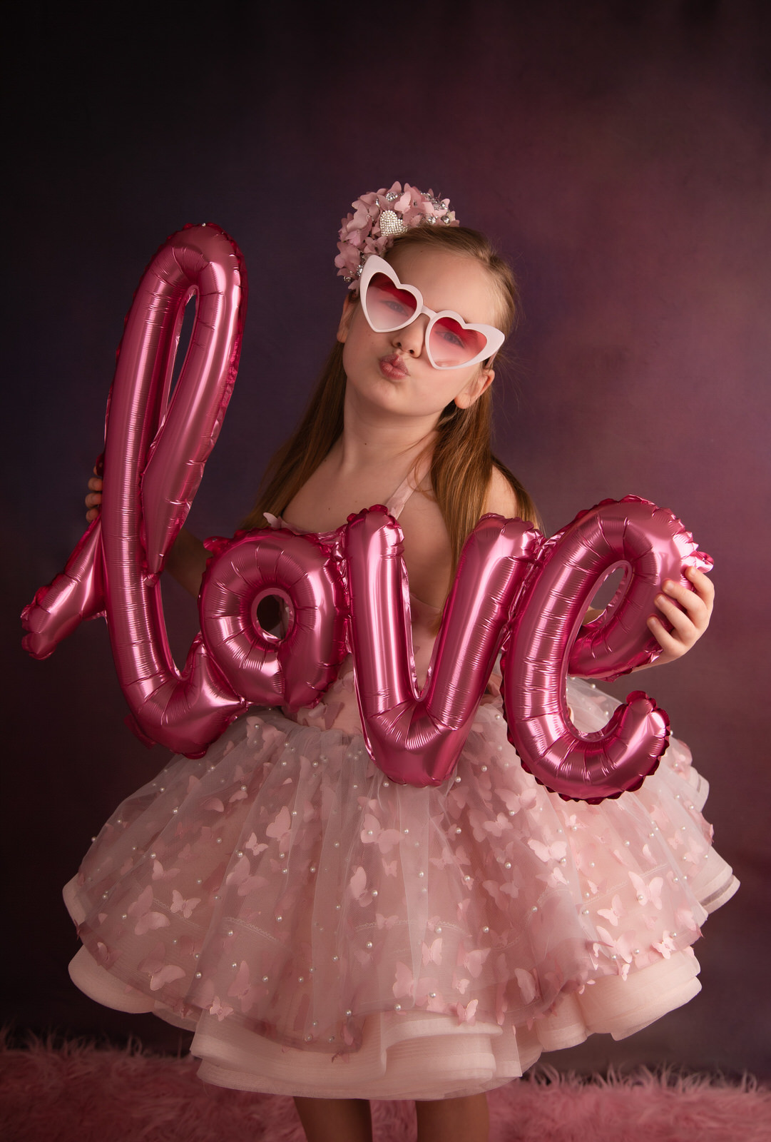 A young girl in a pink tulle tutu dress and heart-shaped sunglasses makes a kiss face with a love balloon fort worth toy stores