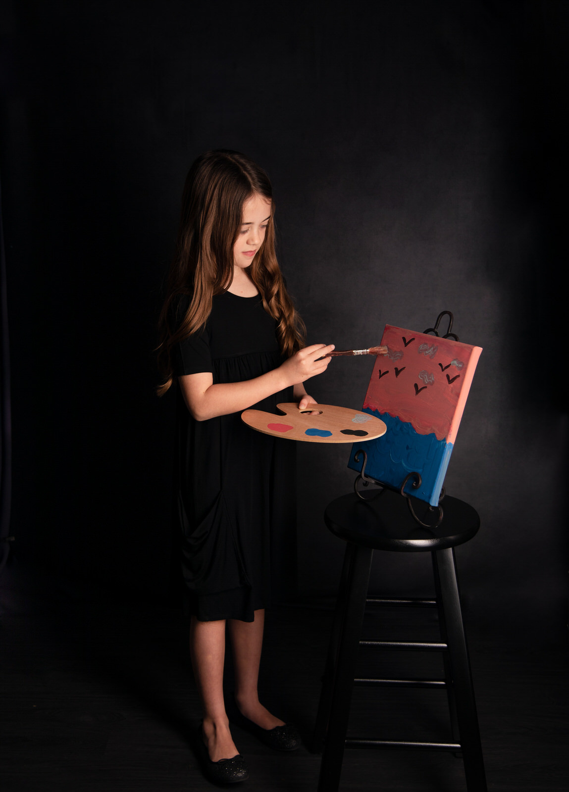 A young girl in a black dress stands in a studio painting an ocean on an easel fort worth toy stores