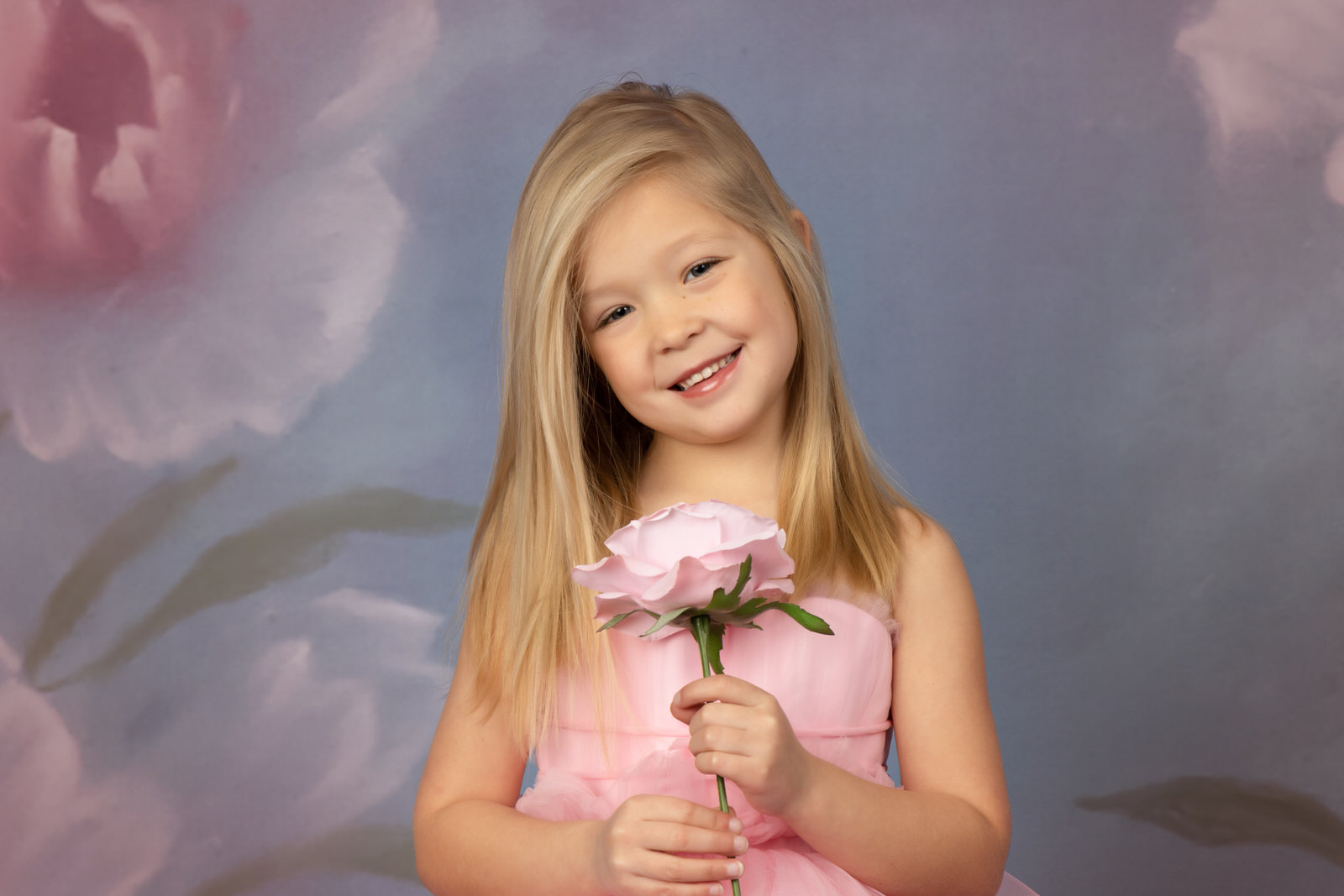 A young girl in a pink dress stands in a studio holding a large pink rose fort worth toy stores
