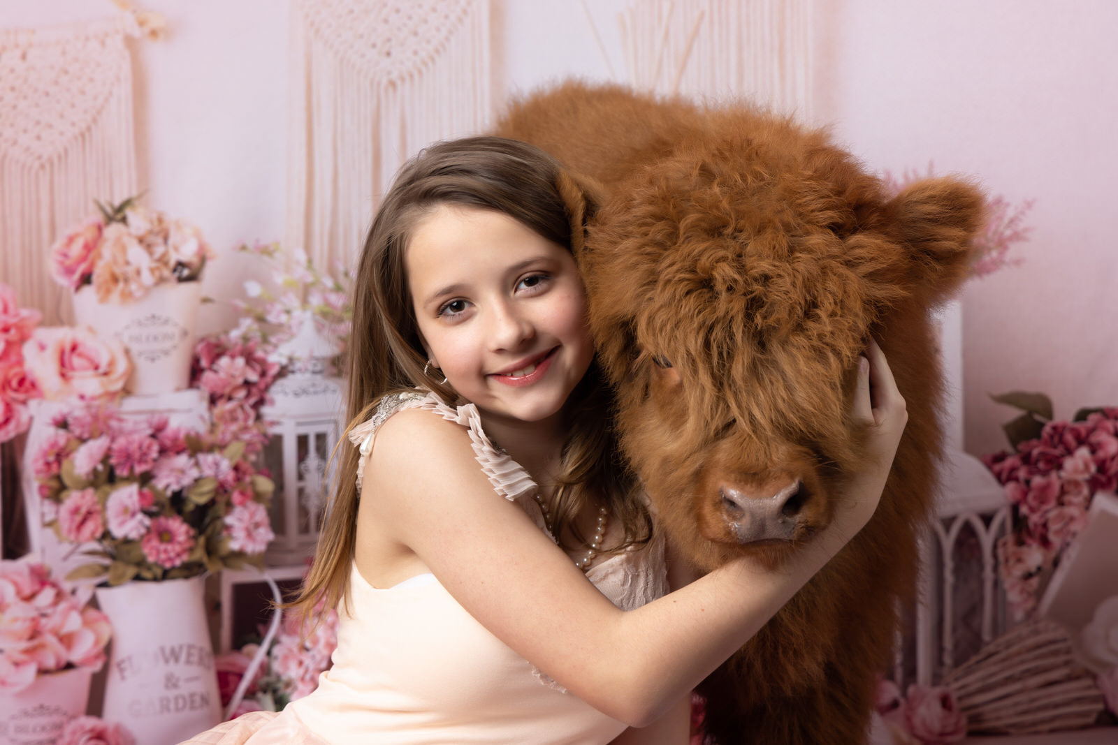 A young girl in a beige dress hugs the face of a fuzzy cow in a studio