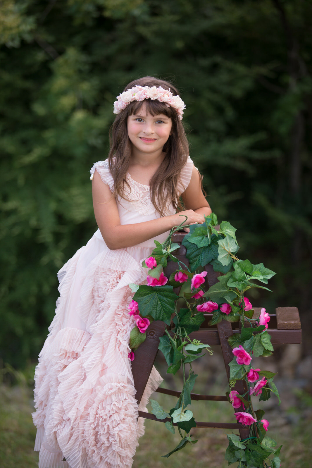 A young girl in a long pink dress leans on a small wooden ladder covered in pink roses