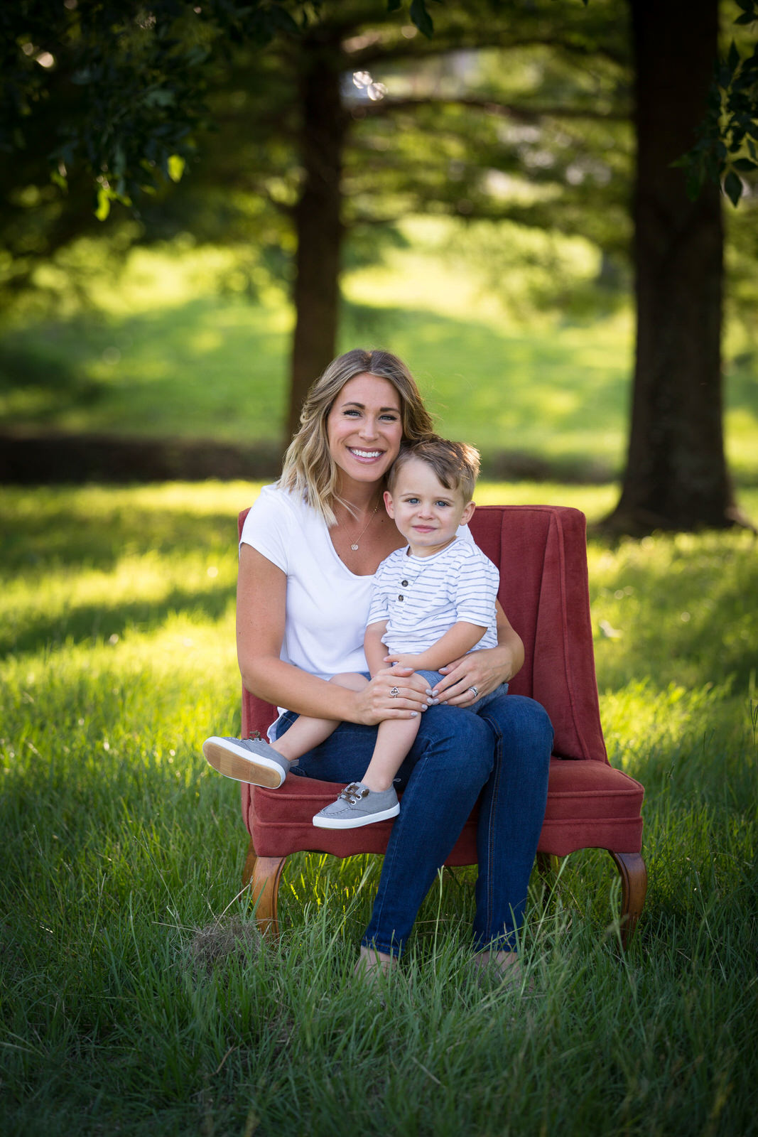 A mother in blue jeans sits in a red chair in a grassy park with her toddler son in her lap midlothian pediatricians