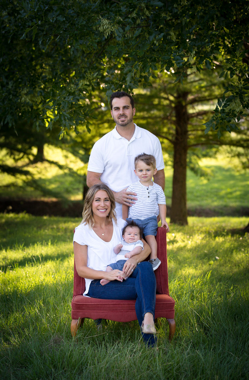 A mother sits in a chair in a park with her newborn son in her lap and husband and toddler son stand behind her midlothian pediatricians