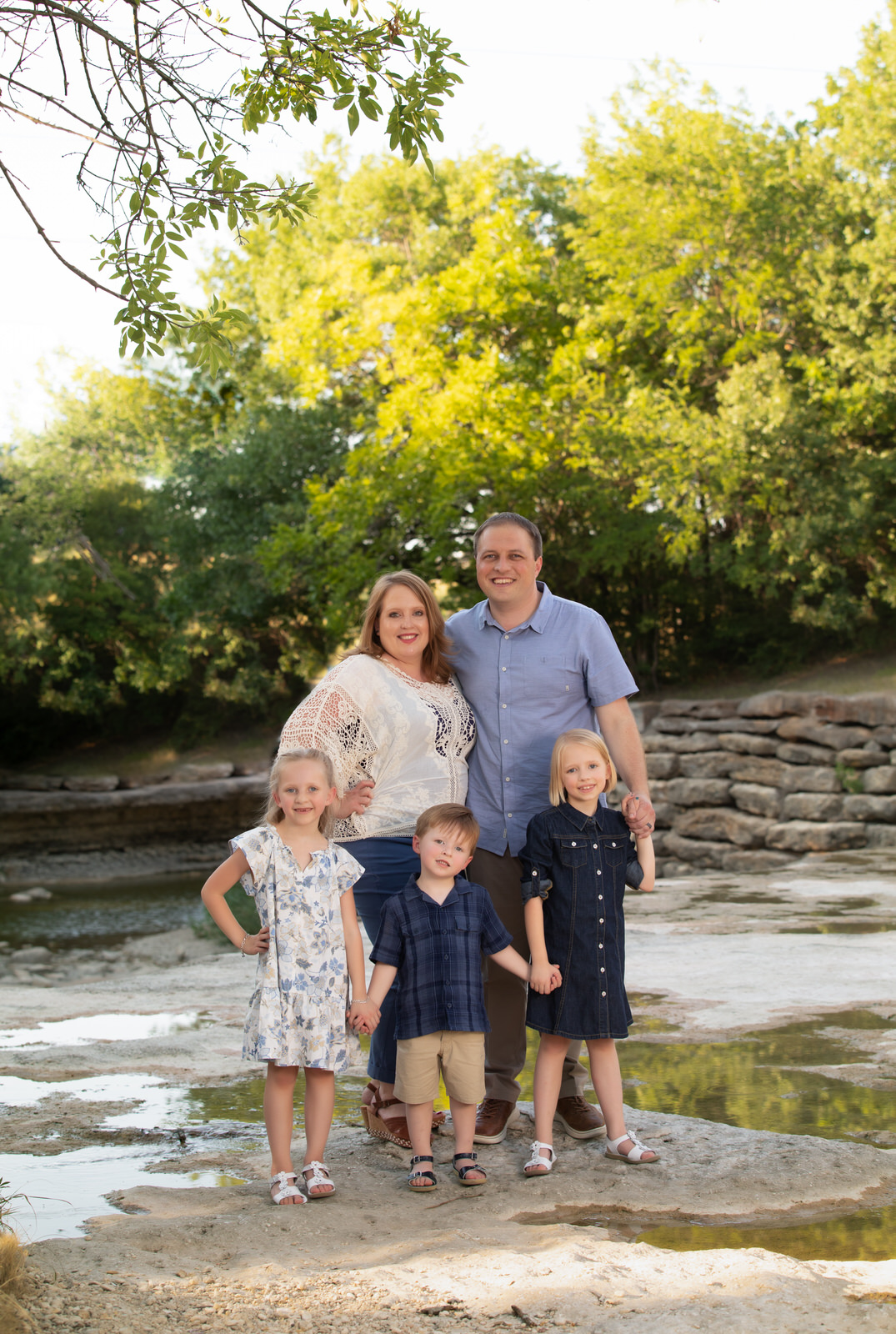 A family of five hold hands while standing on the edge of a river with a stone wall and trees behind them