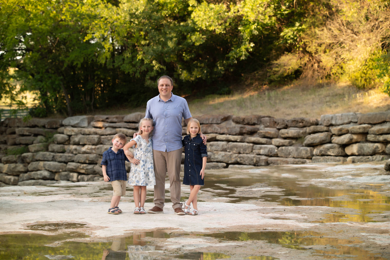A father stands on a river's edge with a stone wall behind him with his three children surrounding him things to do in fort worth with kids