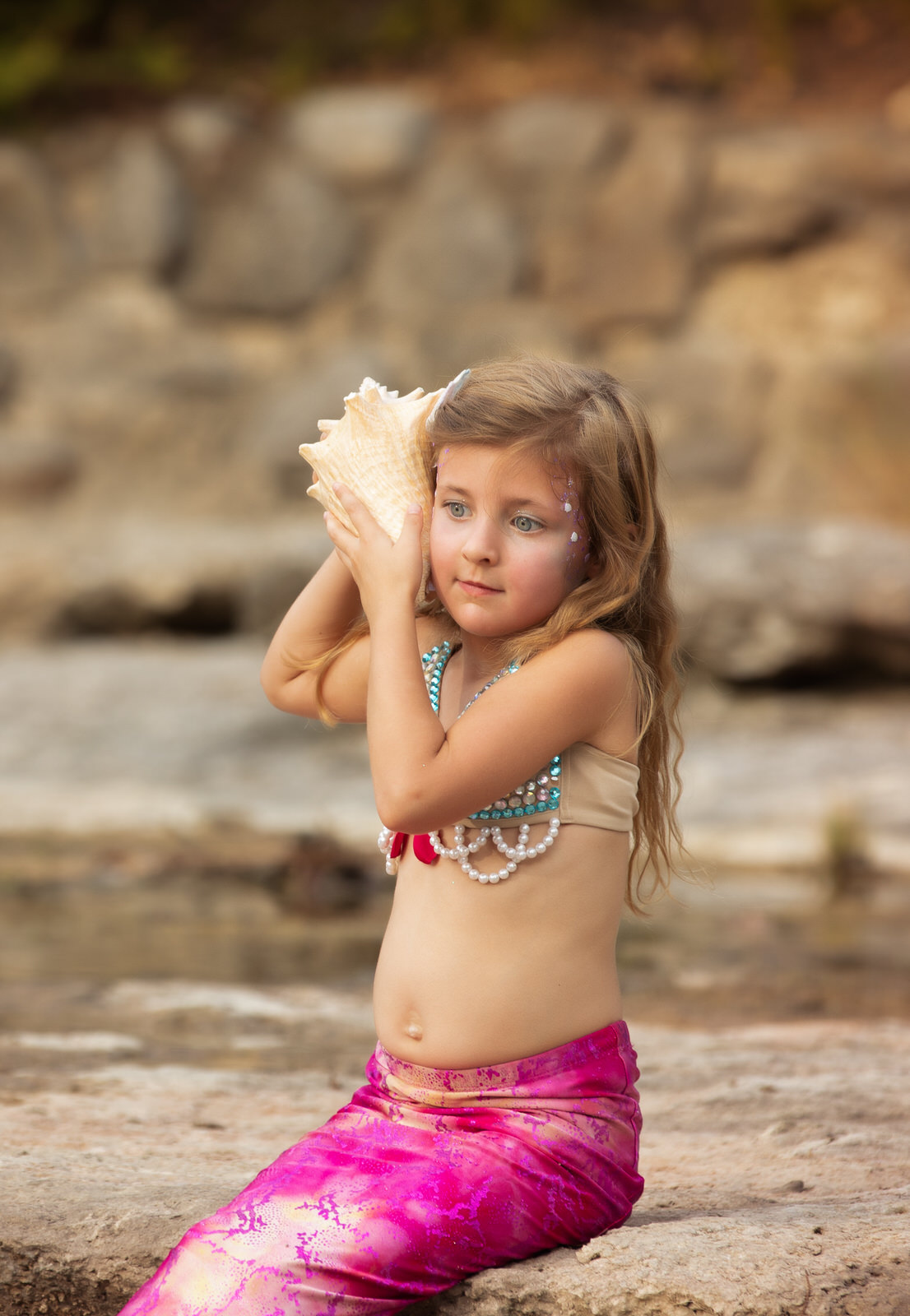 A young girl in a mermaid costume sits on a rock with a conch shell held up to her ear