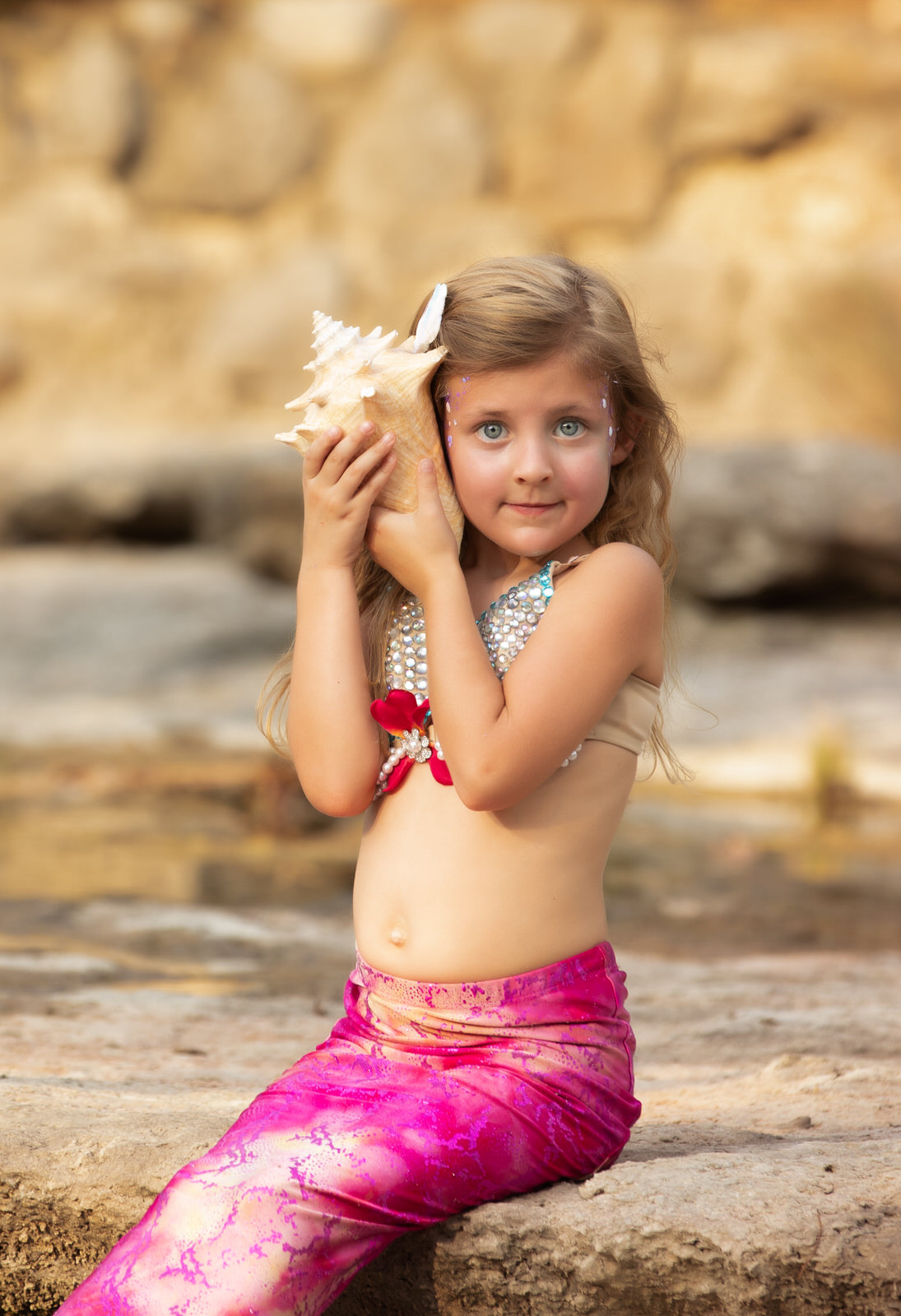 A young girl in a mermaid costume sits on a rock listening to a conch shell