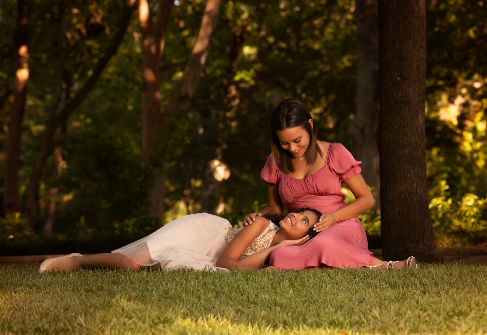 A mother sits in a pink dress in a park with her daughter's head in her lap